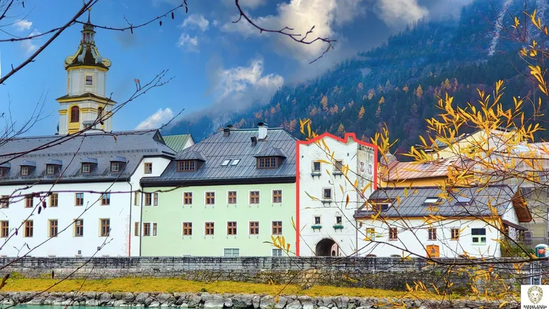 Historische Gebäude entlang eines Flusses mit einem Kirchturm und herbstlicher Berglandschaft im Hintergrund.