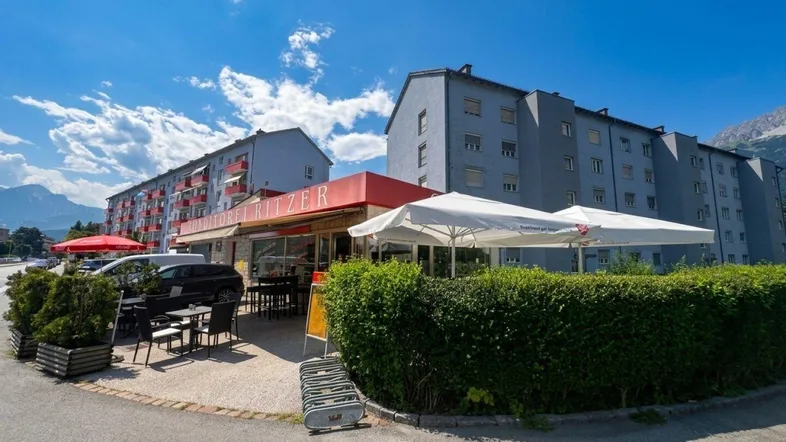 Außenansicht einer Konditorei mit sonniger Terrasse, umgeben von Wohngebäuden und einer Bergkulisse.