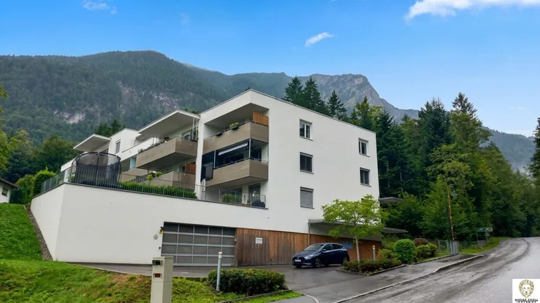 Modernes Apartmentgebäude mit Balkonen und Tiefgarage, umgeben von grünen Bergen.