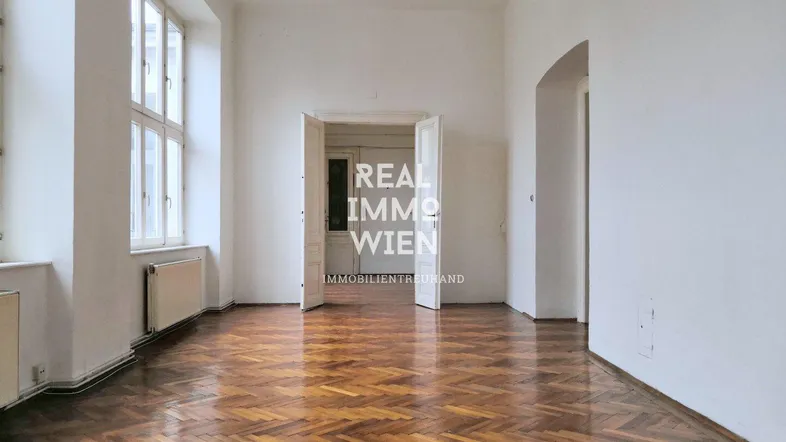 Geräumiges Zimmer mit Fischgrätparkett, hohen Fenstern und weiß gestrichenen Wänden in einem Altbau.