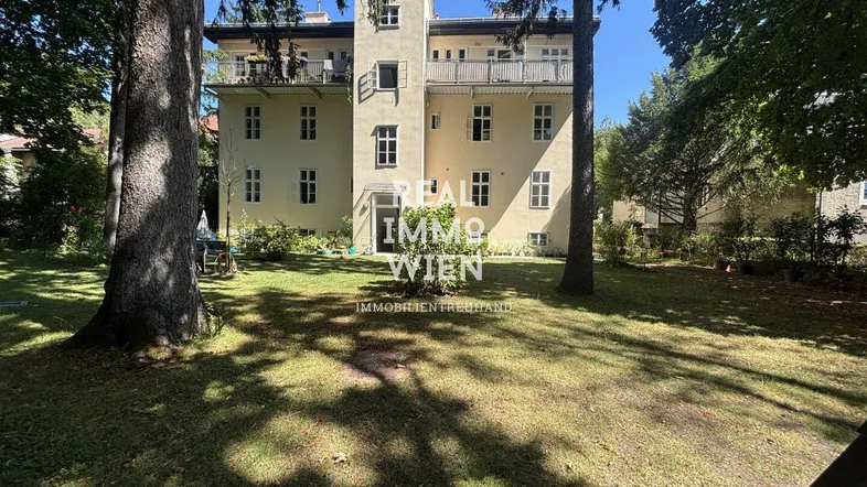 Historisches Gebäude mit heller Fassade und großem, gepflegtem Gartenbereich umgeben von Bäumen.