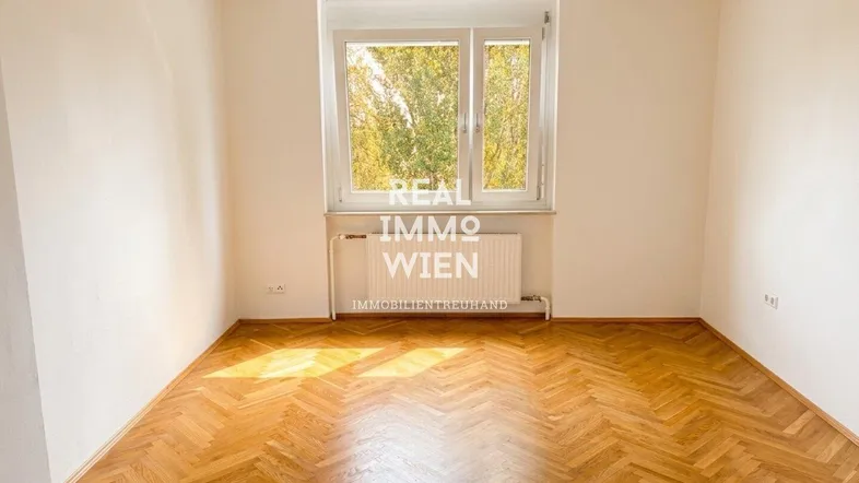 Helles Zimmer mit Fischgrätparkettboden und einem großen Fenster mit Blick ins Grüne.