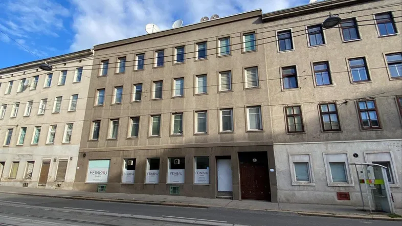 Fassade eines älteren Mehrfamilienhauses an einer Stadtstraße mit Straßenbahnschienen und Satellitenschüsseln auf dem Dach.