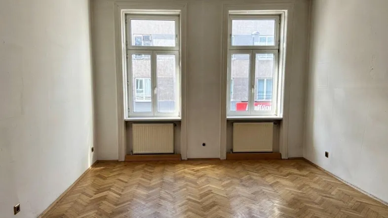 Geräumiges Wohnzimmer mit Fischgrätparkett und zwei großen Fenstern, die viel Tageslicht hereinlassen.