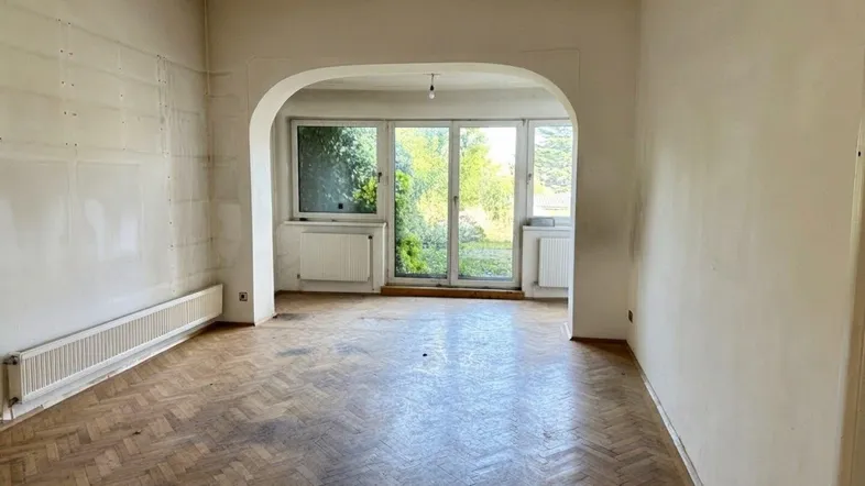 Geräumiges Wohnzimmer mit Fischgrätparkett und großem Durchgang zu einem hellen Bereich mit Fenstern.