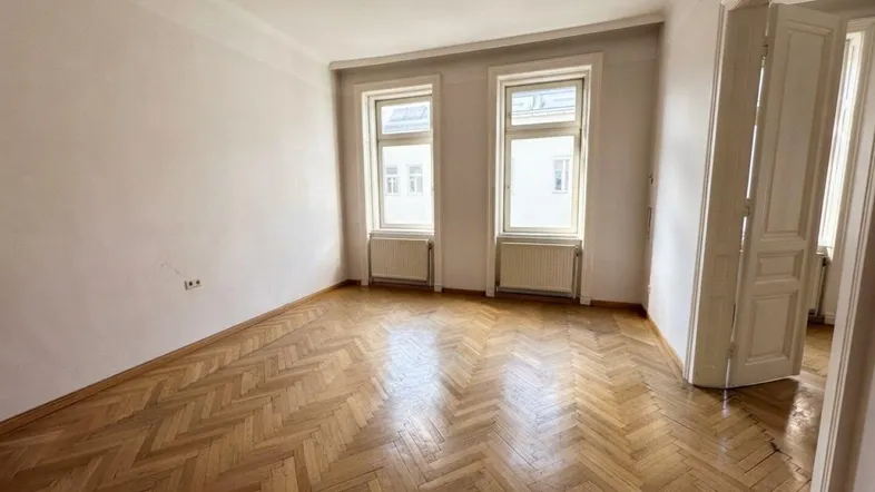 Geräumiges Zimmer mit Fischgrätparkett und zwei großen Fenstern, die viel Tageslicht hereinlassen.