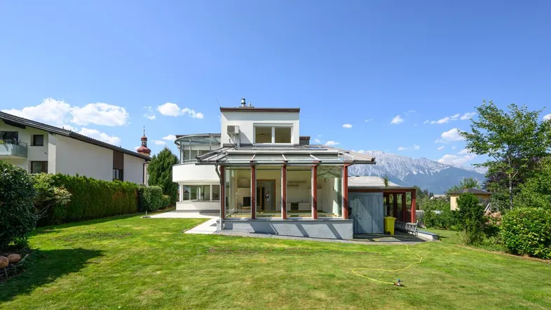 Modernes Haus mit großem Garten, Wintergarten und Bergblick unter blauem Himmel.
