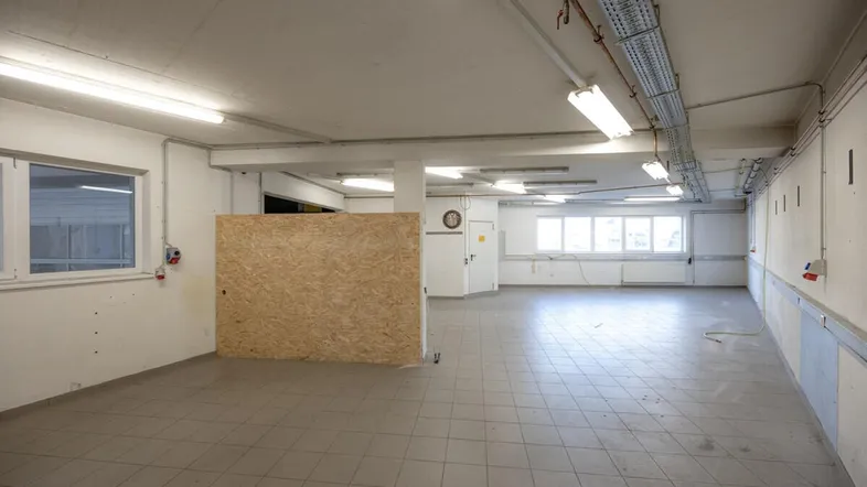 Geräumige, leere Gewerbefläche mit Fliesenboden, großen Fenstern und sichtbaren Installationen an der Decke.