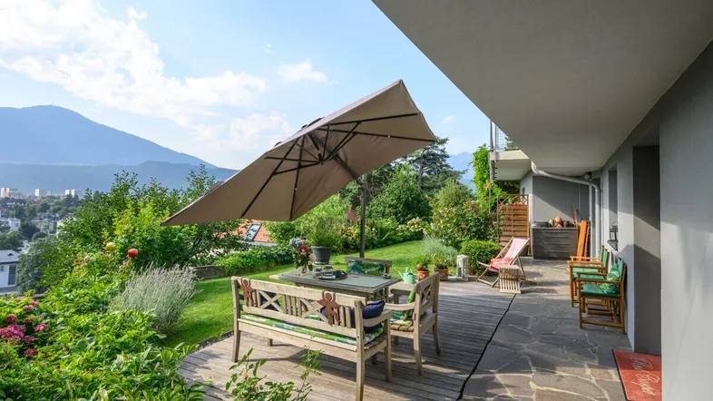 Große Terrasse mit Holzboden, Gartenmöbeln und einem Sonnenschirm, umgeben von üppigem Grün und Bergblick.
