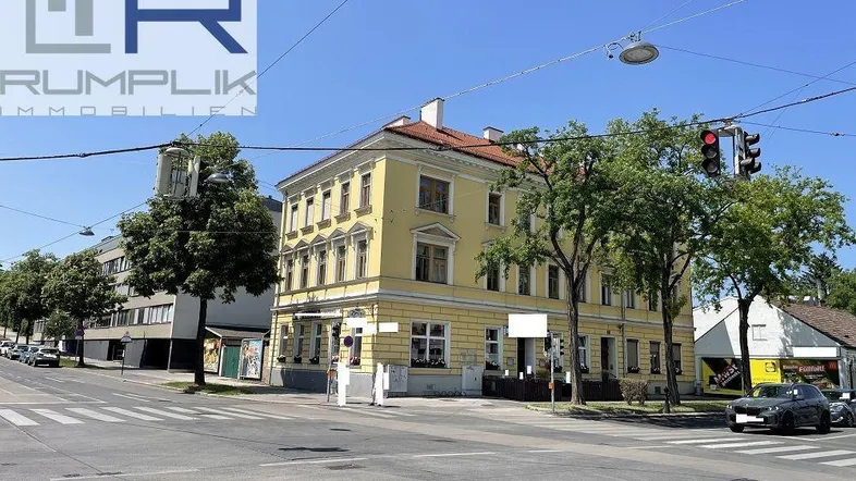Gelbes Eckgebäude an einer belebten Kreuzung mit Bäumen und klarem Himmel.