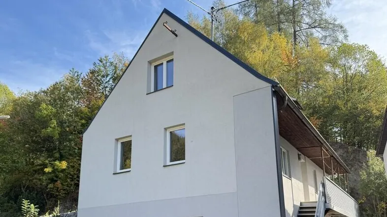 Helles Haus mit Giebeldach und Außentreppe, umgeben von Bäumen unter blauem Himmel.