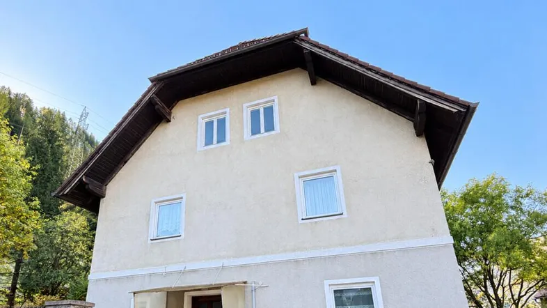 Hausfassade mit beiger Putzfassade, Satteldach und mehreren Fenstern, umgeben von Bäumen.