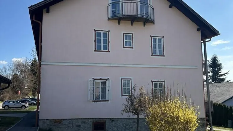Seitenansicht eines Wohnhauses mit hellrosa Fassade, Satteldach und kleinem Balkon im Obergeschoss.