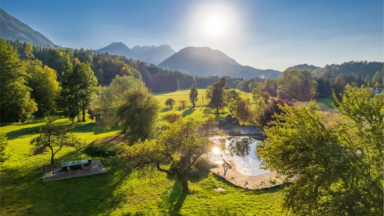 Sonnige Landschaft mit einem natürlichen Teich, umgeben von grünen Wiesen und majestätischen Bergen im Hintergrund.