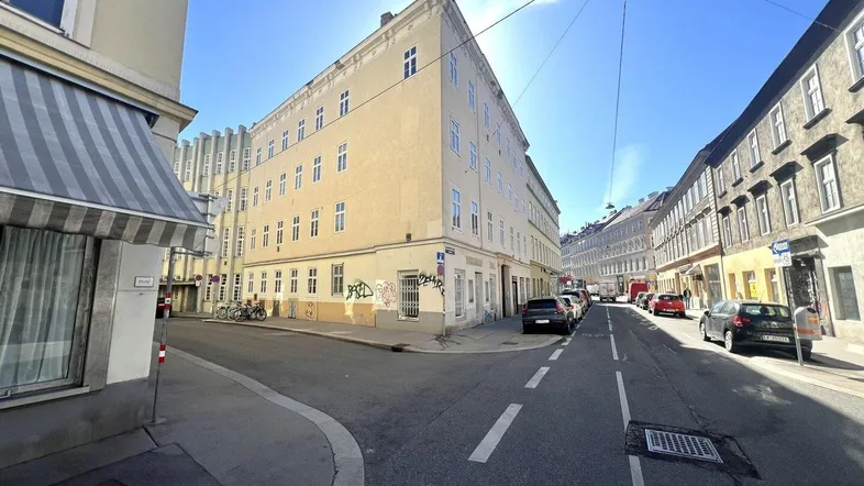 Blick auf ein Eckgebäude in einer städtischen Umgebung mit geparkten Autos und blauem Himmel.