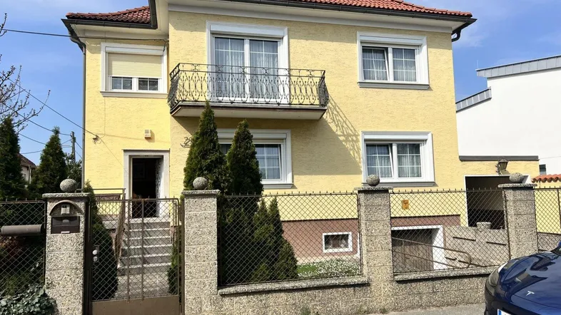 Gelbes Einfamilienhaus mit rotem Ziegeldach, kleinem Balkon und umzäuntem Vorgarten.