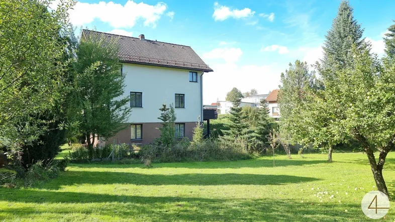 Großer Gartenbereich mit Rasenfläche und Bäumen, das Haus im Hintergrund unter blauem Himmel.