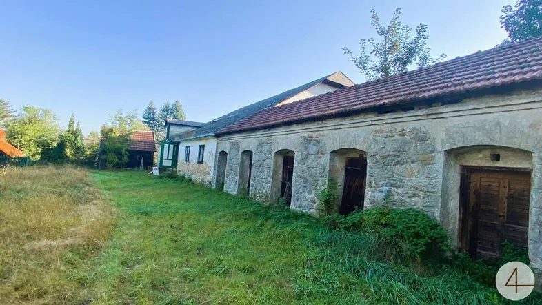 Lange Fassade eines alten Gebäudes mit Steinbögen und einer weiten, ungemähten Wiese davor unter blauem Himmel.