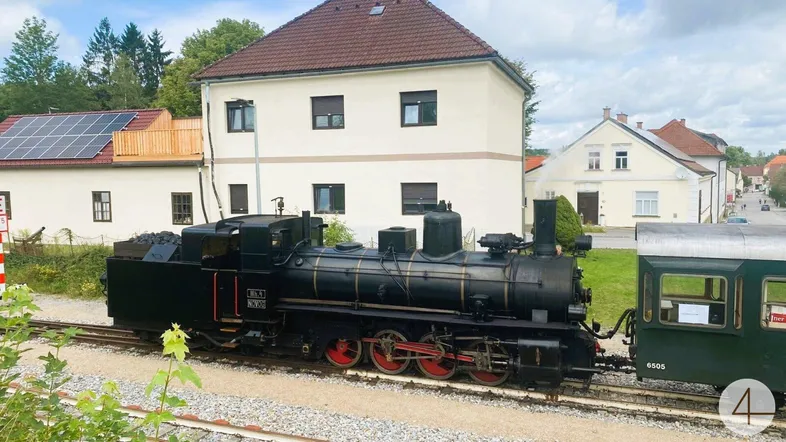 Historische Dampflokomotive auf Gleisen vor ländlichen Gebäuden mit Solaranlage auf dem Dach.