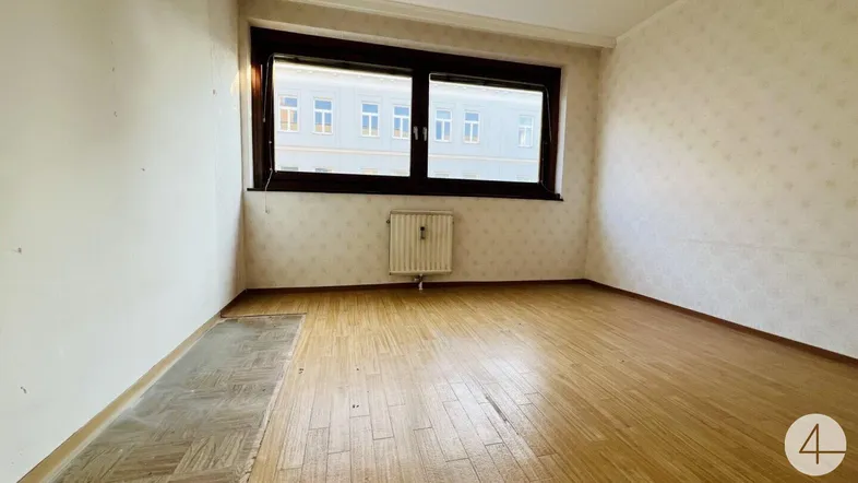 Helles Zimmer mit Holzfußboden und Blick auf die umliegenden Gebäude, mit älterer Tapete.