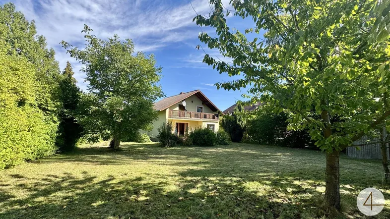 Haus mit großem Garten und Balkon, umgeben von viel Grün unter blauem Himmel.