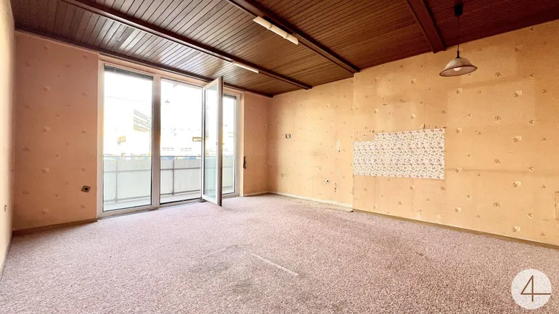 Geräumiger Wohnraum mit Teppichboden, Holzdecke und Zugang zum Balkon, renovierungsbedürftig.