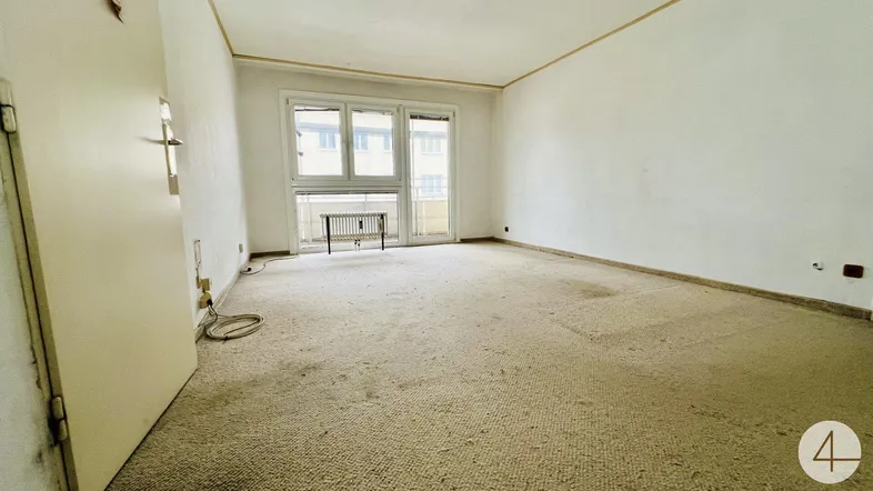 Leeres Wohnzimmer mit altem Teppichboden und Zugang zu einem kleinen Balkon.