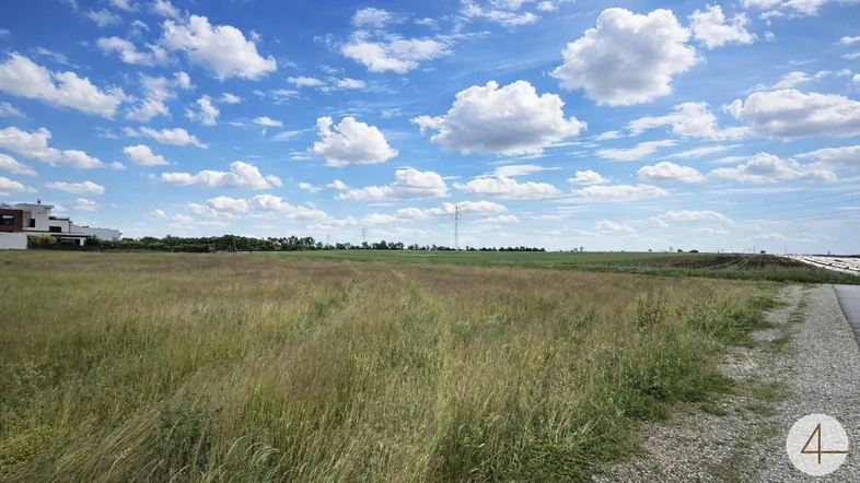 Weitläufiges Grundstück mit grüner Wiese unter blauem Himmel und vereinzelten Häusern im Hintergrund.