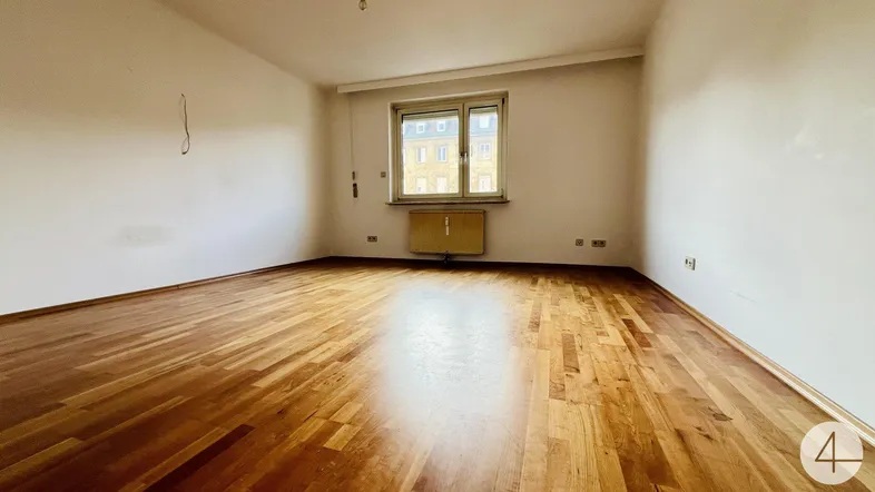Helles, leeres Wohnzimmer mit glänzendem Parkettboden und einem Fenster mit Blick nach draußen.