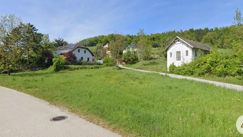 Blick auf ein grünes Grundstück mit umliegenden Häusern und bewaldeten Hügeln im Hintergrund.