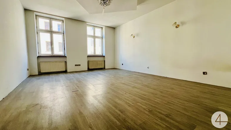 Heller, leerer Raum mit Holzfußboden, zwei Fenstern und hohen Decken.