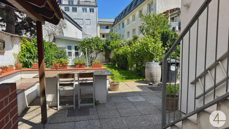 Gepflasterter Gartenbereich mit Grünfläche, Gartenhaus und einer kleinen Außenbar, umgeben von Gebäuden.