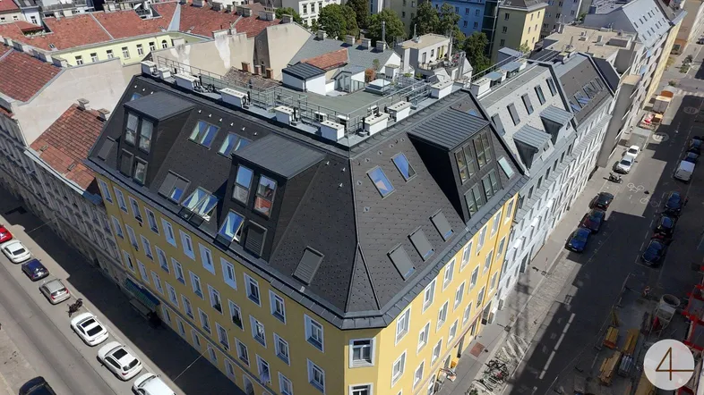 Luftaufnahme eines Eckgebäudes mit gelber Fassade und modernem Mansarddach in städtischer Umgebung.