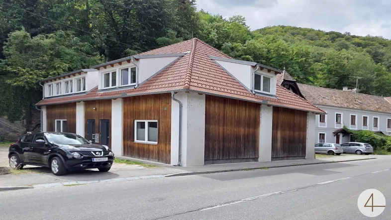Modernes Gebäude mit Holzfassade und Satteldach, umgeben von grünen Hügeln und Bäumen.