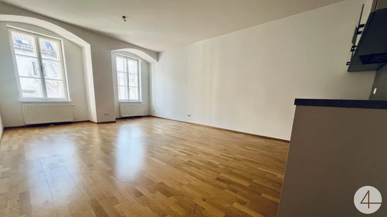 Helles Wohnzimmer mit Holzboden und zwei Fenstern, die viel Tageslicht hereinlassen.