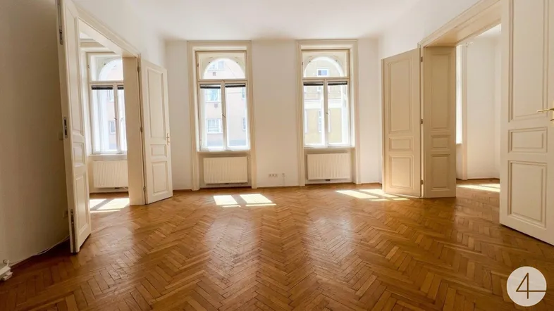 Helles Zimmer mit Fischgrätparkett, hohen Fenstern und mehreren Türen, ideal als Wohnbereich.