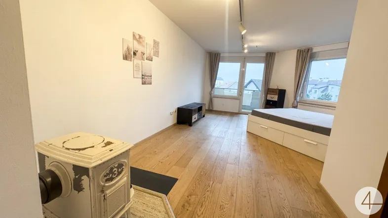 Helles Zimmer mit Holzboden, einem Ofen und Zugang zu einem Balkon, ideal als Wohn- und Schlafbereich.