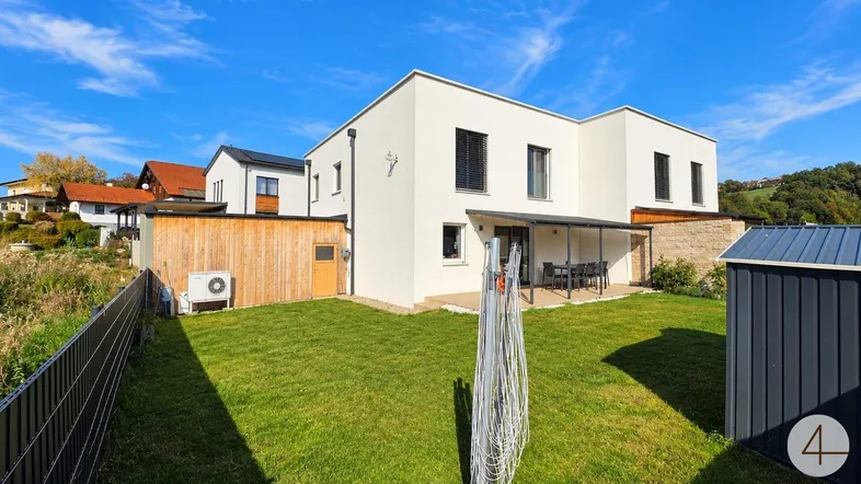 Rückansicht eines modernen Hauses mit großem Garten, Terrasse und Geräteschuppen unter blauem Himmel.
