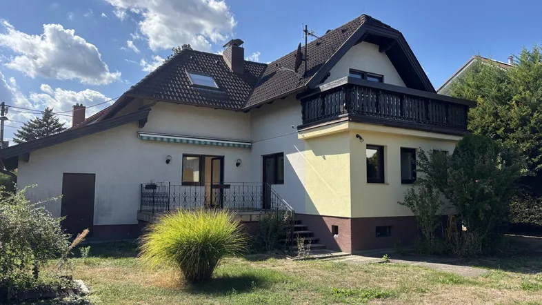 Hausfassade mit Gartenbereich und Balkon unter blauem Himmel, umgeben von Grün.