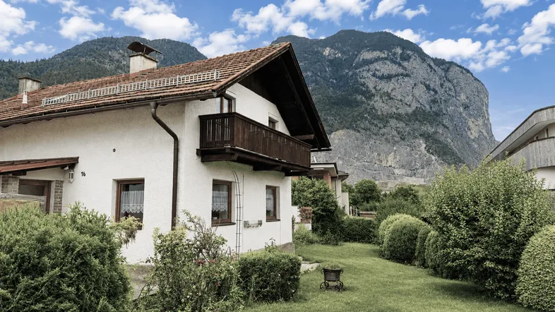 Weißes Haus mit Holzbalkon, gepflegtem Garten und beeindruckendem Bergblick im Hintergrund.