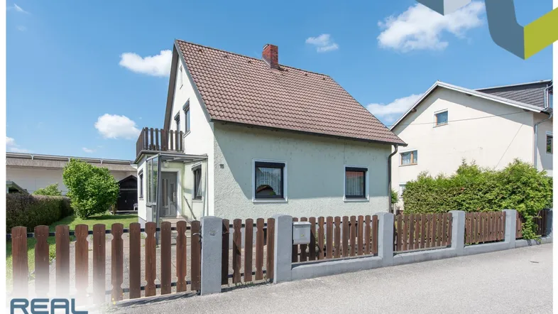 Einfamilienhaus mit Satteldach, kleinem Vorgarten und Holzzaun unter blauem Himmel.