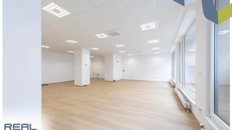 Geräumiger, heller Büroraum mit großen Fenstern, hellem Laminatboden und moderner Deckenbeleuchtung.