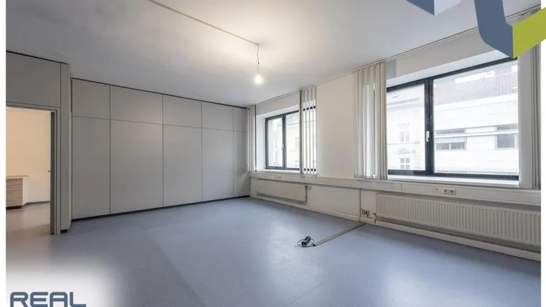 Geräumiger Büroraum mit großen Fenstern, hellen Wänden und grauem Bodenbelag.