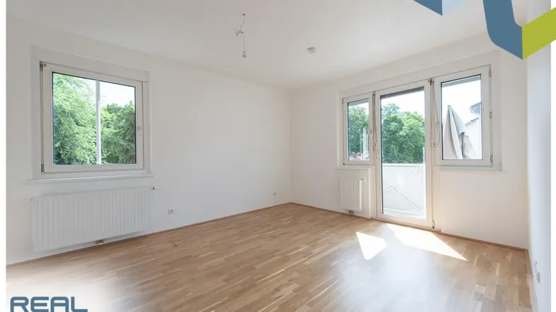 Helles Zimmer mit Parkettboden, weißen Wänden und Zugang zu einem kleinen Balkon.