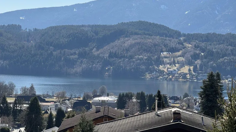 Panoramablick auf den See und die umliegenden Berge mit Häusern im Vordergrund unter blauem Himmel.