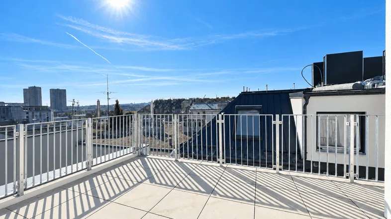 Sonnige Dachterrasse mit weitem Blick über die Stadt und umliegende Gebäude unter blauem Himmel.