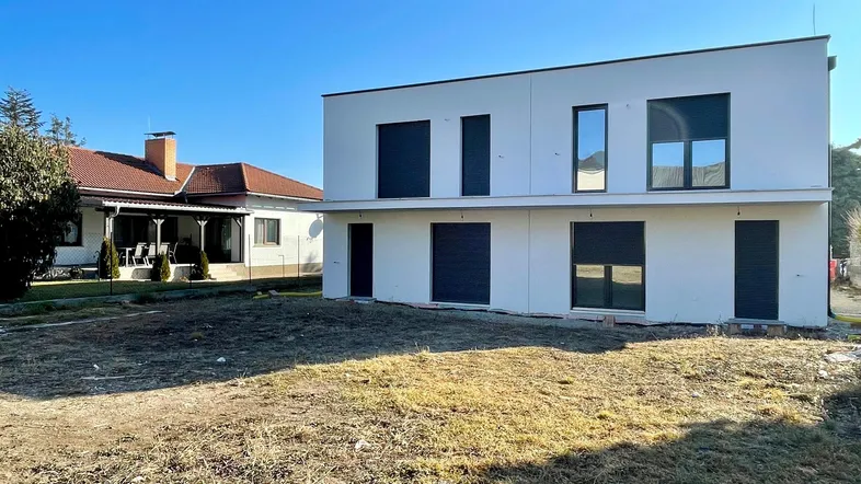 Modernes, weißes Neubauhaus mit dunklen Fensterläden und unbebautem Vorgarten.