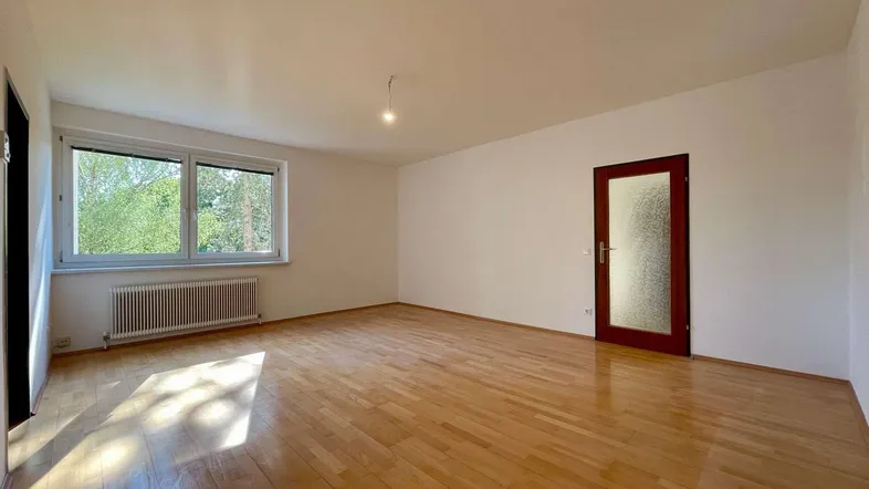 Helles, leeres Zimmer mit Parkettboden und einem Fenster, das viel Tageslicht hereinlässt.