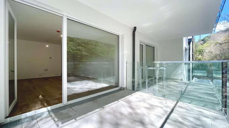 Heller Balkon mit Glasgeländer und Zugang zu einem leeren Raum mit Holzboden, umgeben von Natur.