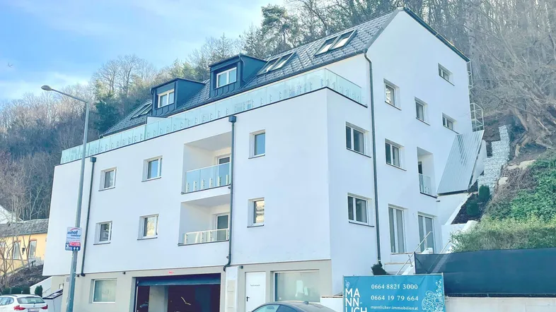 Modernes Mehrfamilienhaus mit weißer Fassade, Balkonen und Dachgauben in grüner Hanglage.